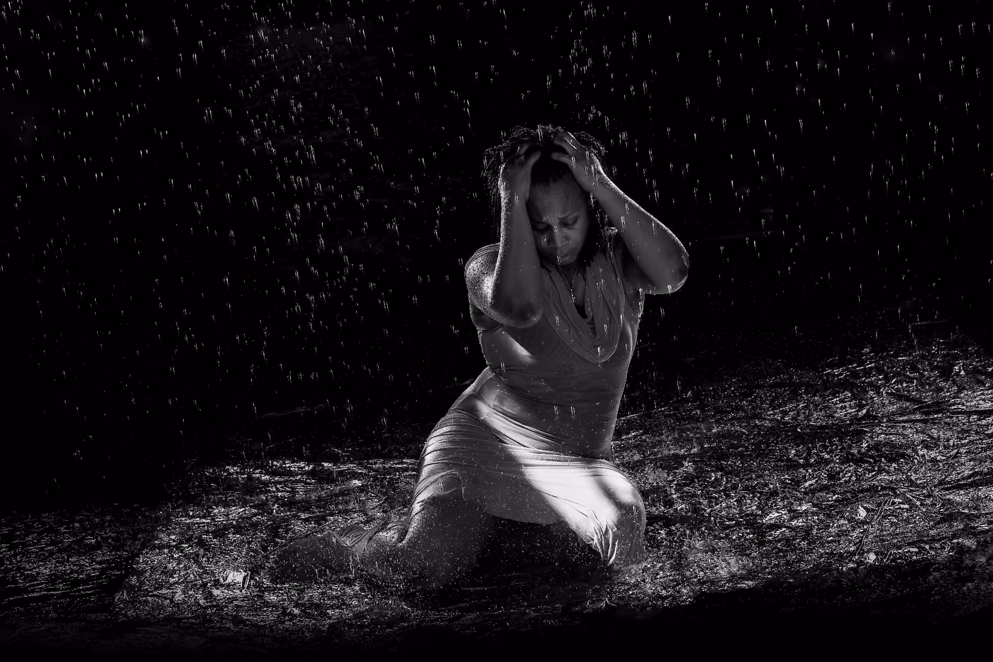 Woman kneeling under falling water in a dark room, white slip